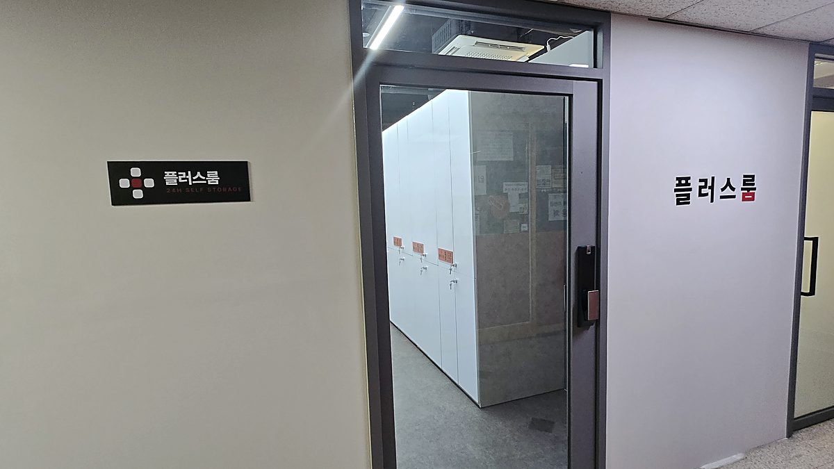 플러스룸 입구 — 벽면 명판과 플러스룸 레터링, 유리문 너머로 보이는 보관 유닛