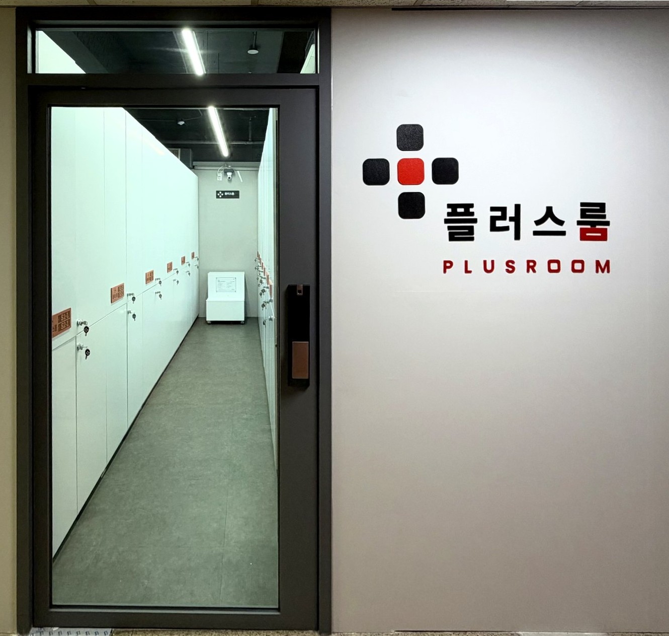 플러스룸 구의동 셀프스토리지 주출입문 정면 — 벽면의 플러스룸 입체 로고 사인과 유리문 너머로 이어지는 보관 유닛 통로